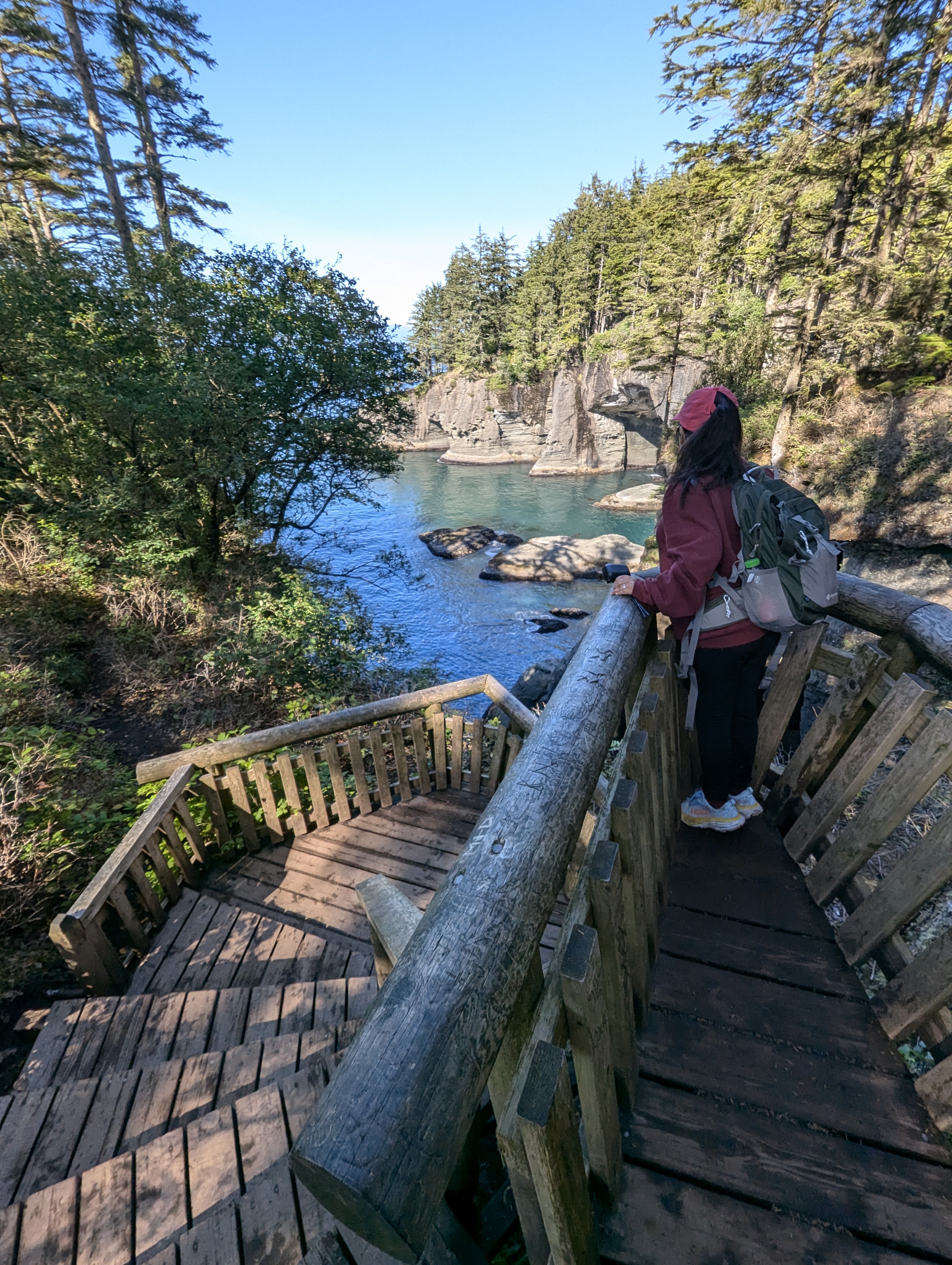 Petite young woman with black hair, Minerva Barrios, wearing a red jacket and backpack, stands on a wooden viewing platform at Cape Flattery, looking out over the ocean for whales, surrounded by coastal cliffs, evergreen trees, and clear blue skies