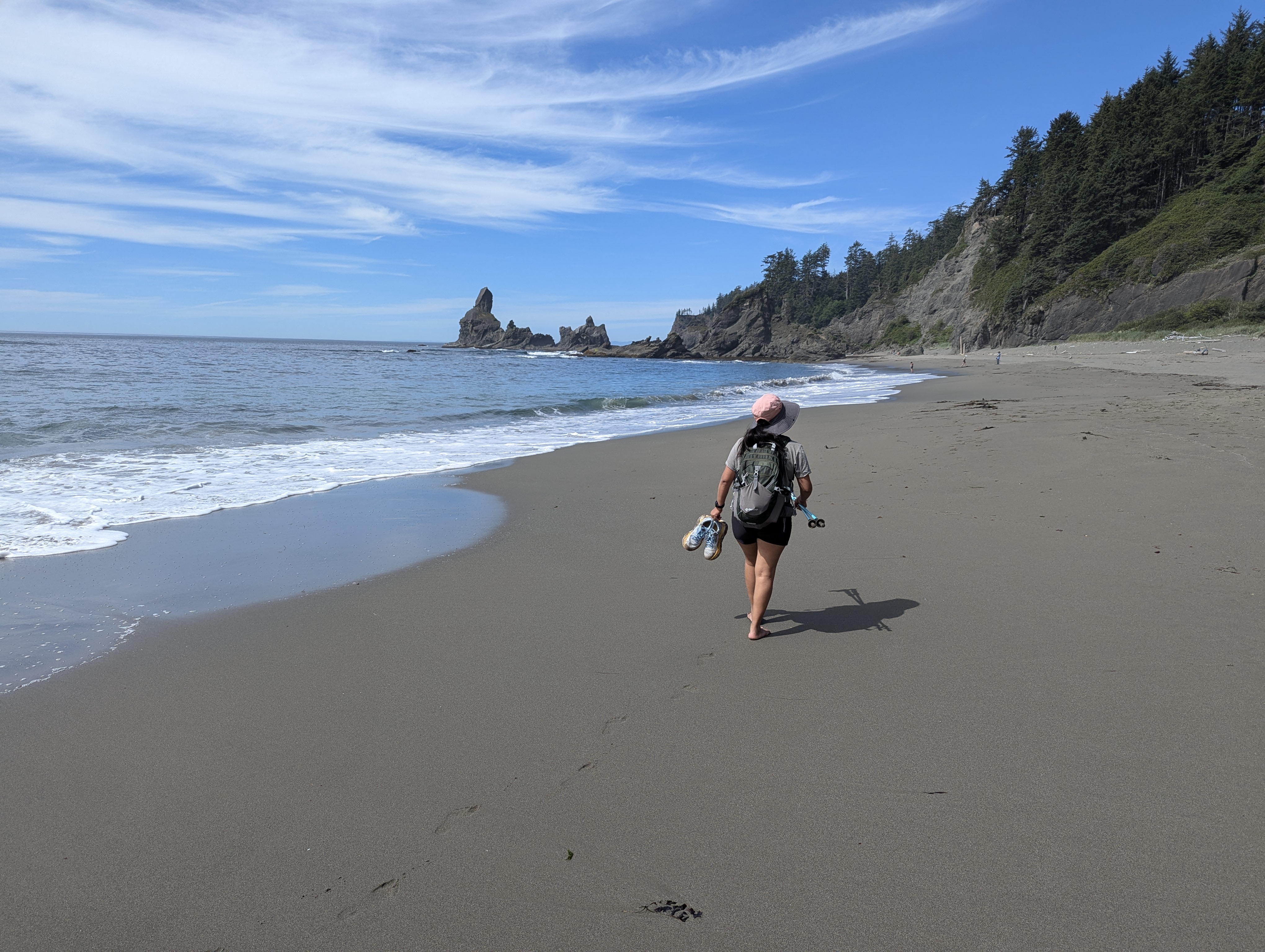 Petite young woman with black hair, Minerva Barrios, hiking on Shi-Shi Beach in Olympic National Park, walking barefoot across soft sand toward sea stacks and rocky cliffs under a sunny blue sky streaked with clouds.