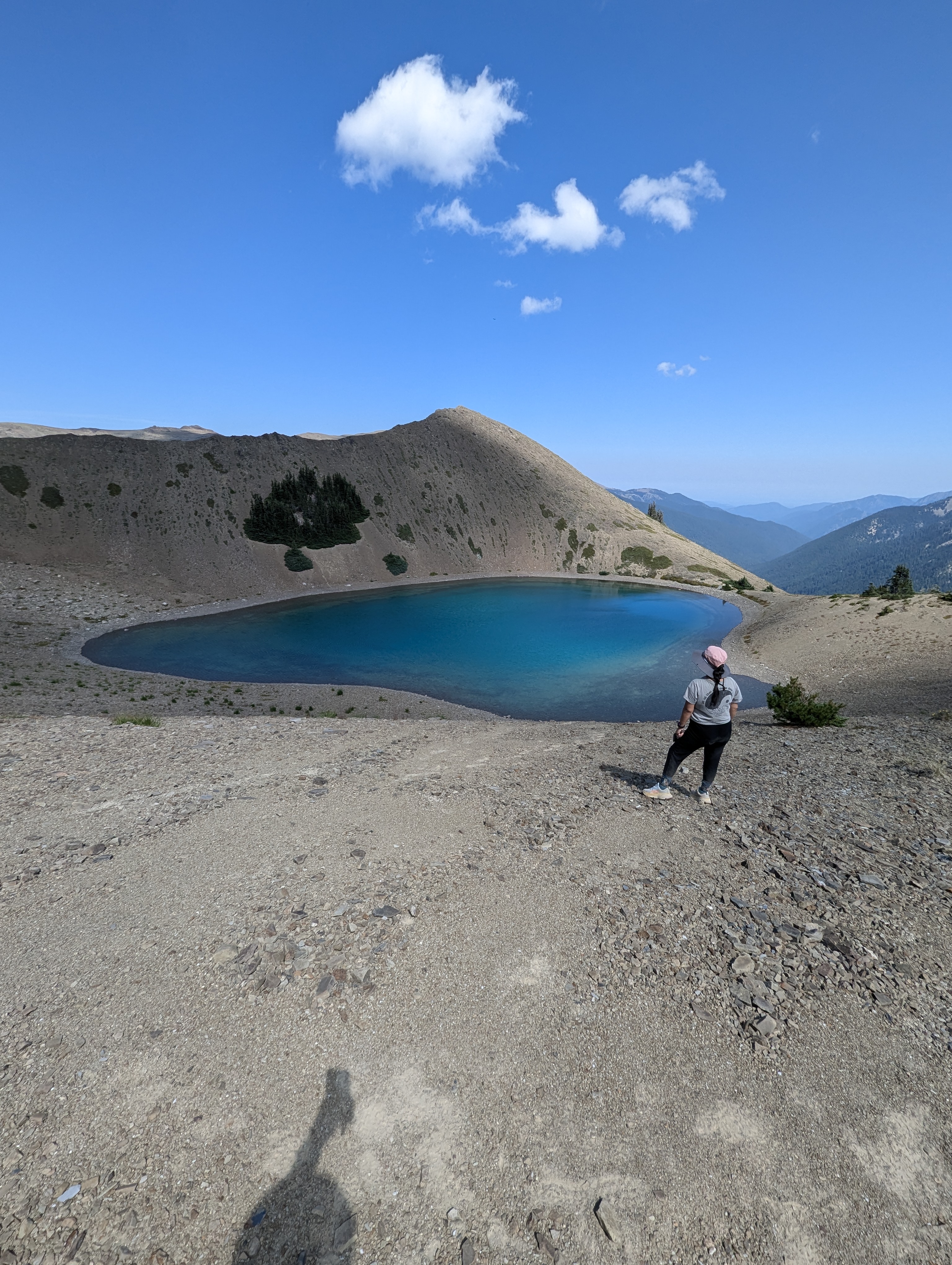Petite young woman with black hair, Minerva Barrios, hiking Grand Lake Trail in Olympic National Park, standing near a turquoise alpine lake beneath a bright blue sky with scattered clouds, surrounded by rugged mountain views