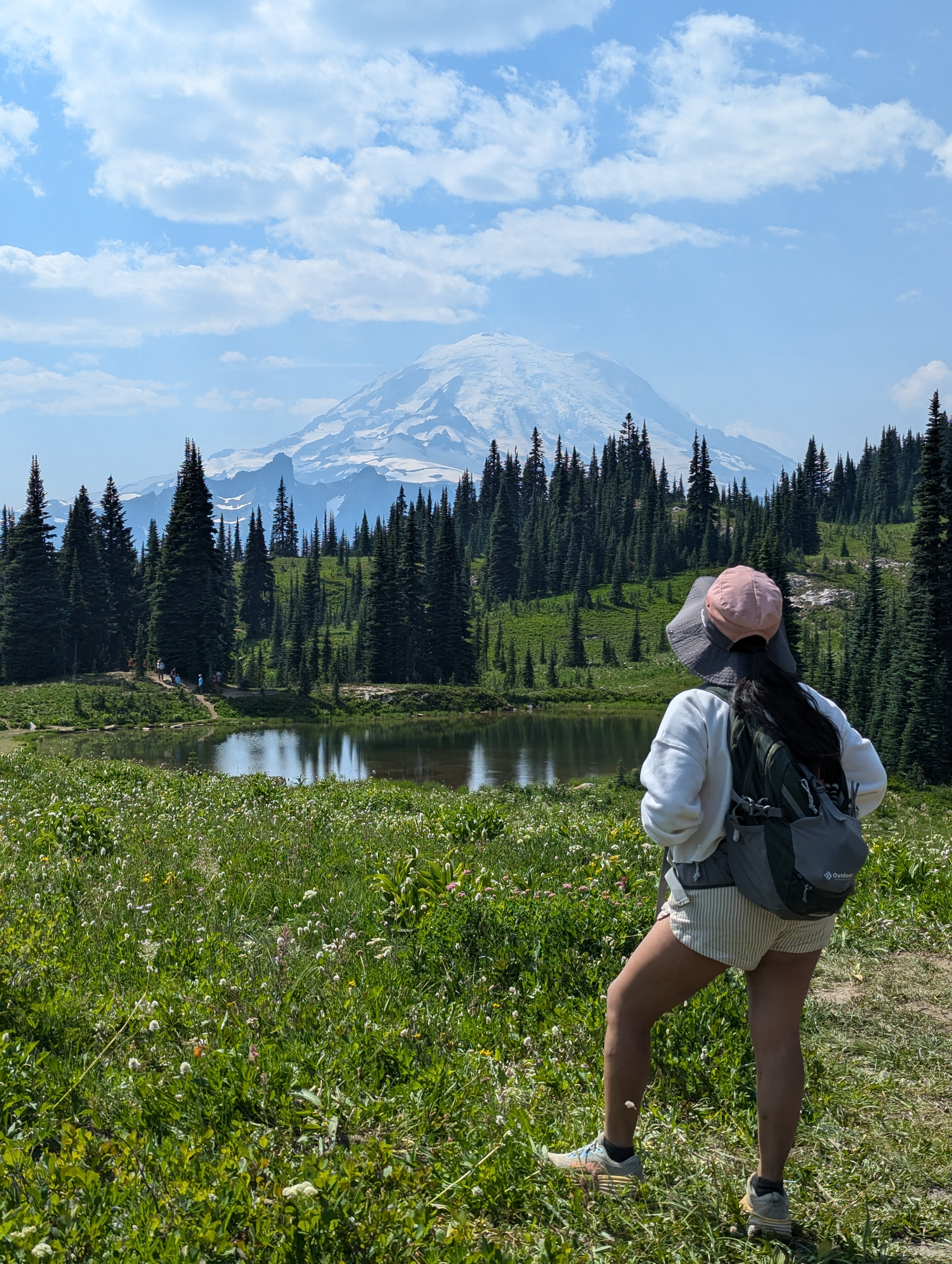Minerva Barrios, a petite young woman with long black hair in a pink cap and backpack, standing in a lush green meadow full of wildflowers, admiring the towering snow-capped Mount Rainier beyond a small reflective pond and evergreen trees.