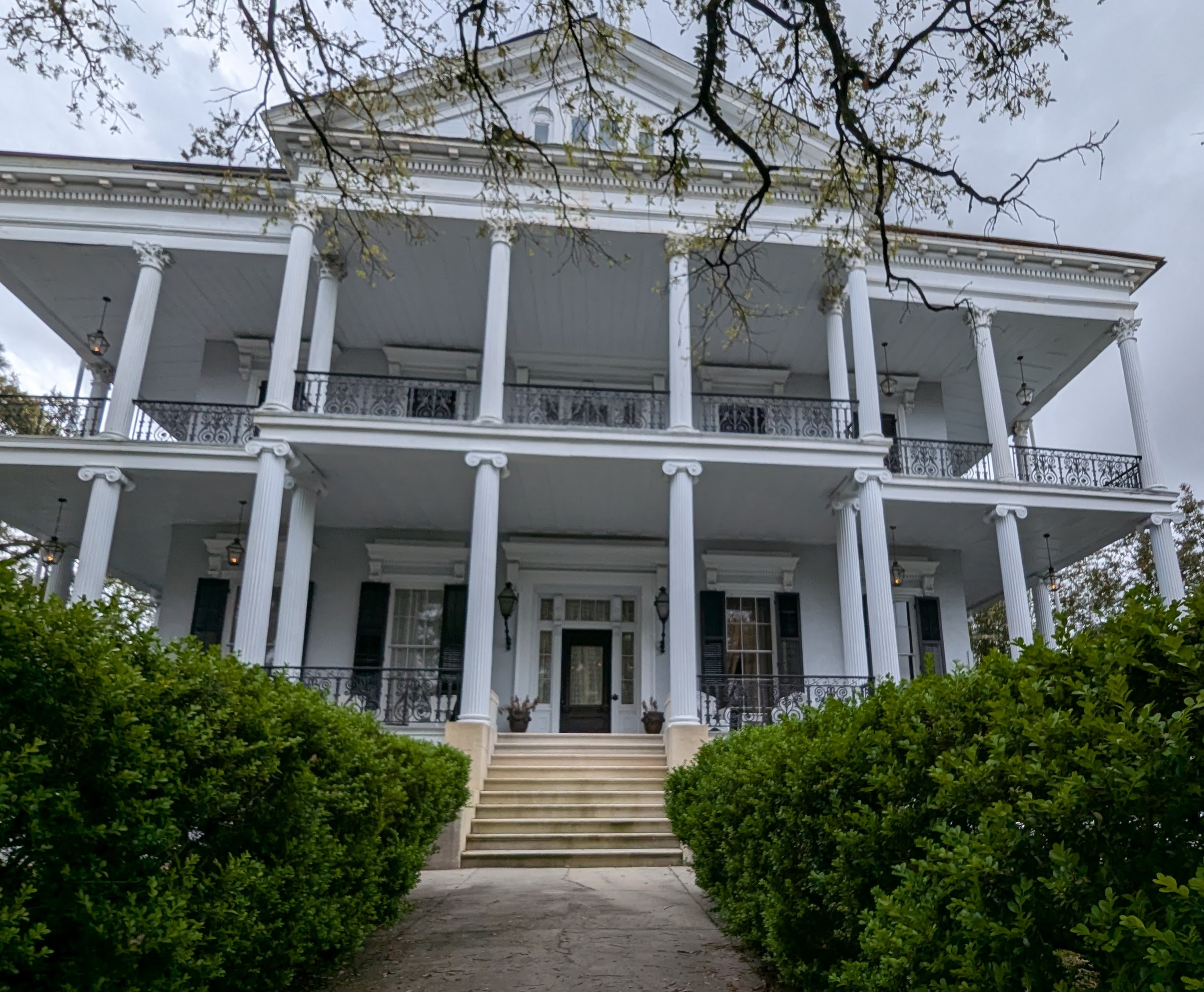 Front view of Buckner Mansion, a grand white Greek Revival-style home in New Orleans with tall columns, ironwork balconies, and a staircase leading to a central double door, framed by manicured hedges.