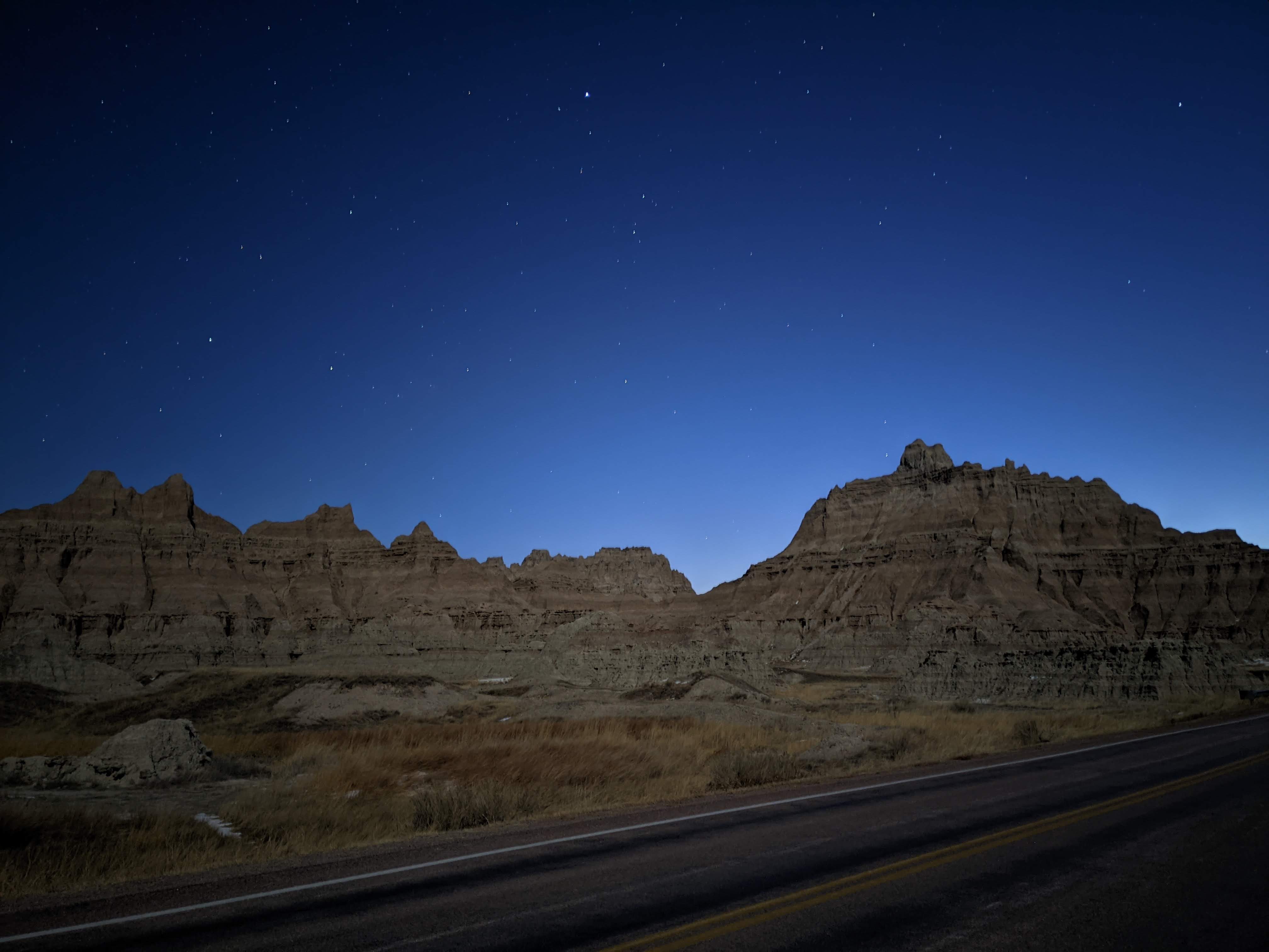 A starry night sky casts a tranquil glow over the jagged rock formations of Badlands National Park, with a quiet road in the foreground leading into the rugged landscape. The scene captures the park’s stark beauty and remote stillness under the twilight.
