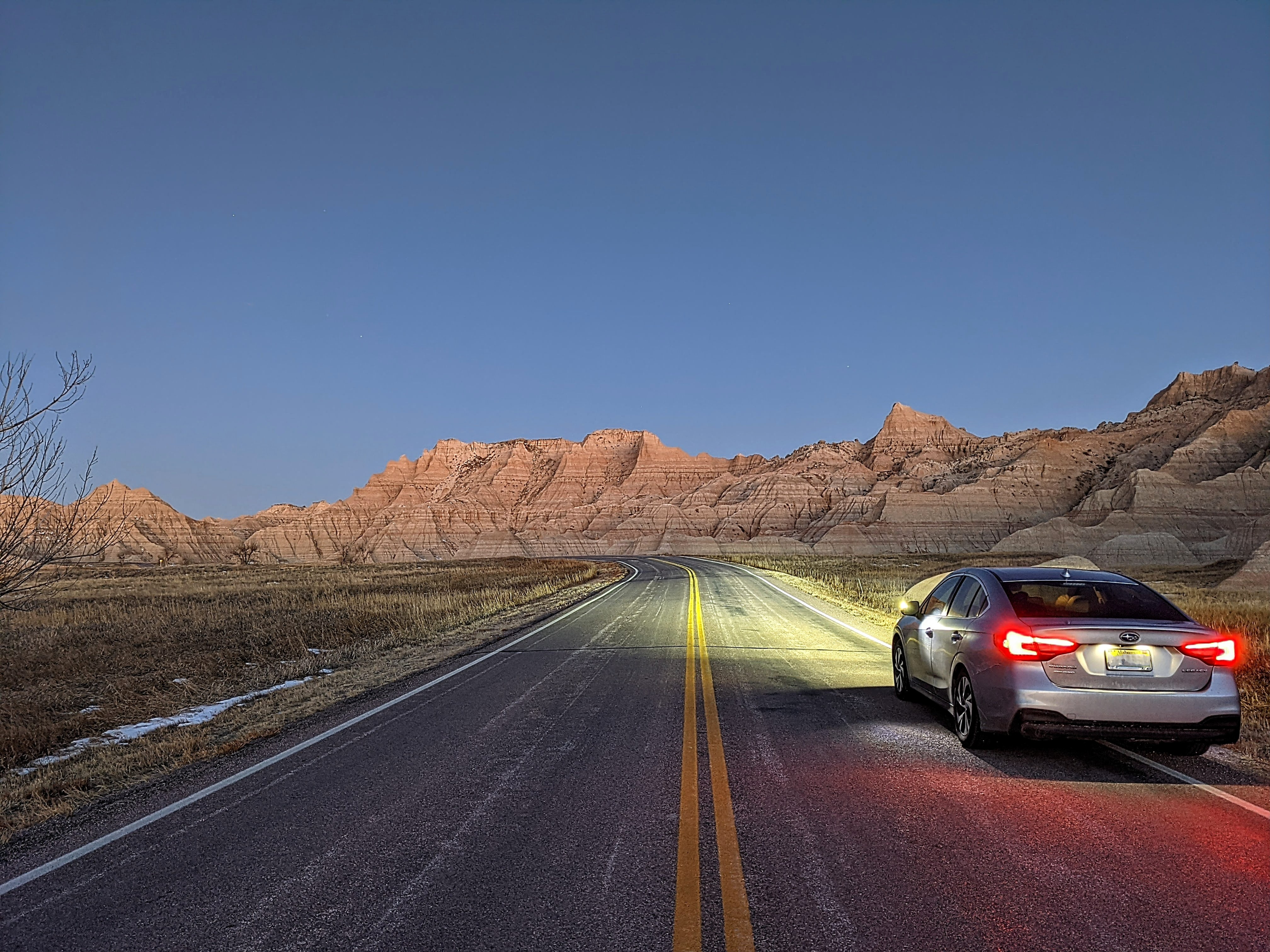 A silver car is parked on the roadside at dawn in Badlands National Park, its headlights illuminating the empty highway ahead. The rugged, layered rock formations of the park glow faintly in the early light under a clear blue sky, creating a serene and dramatic landscape.