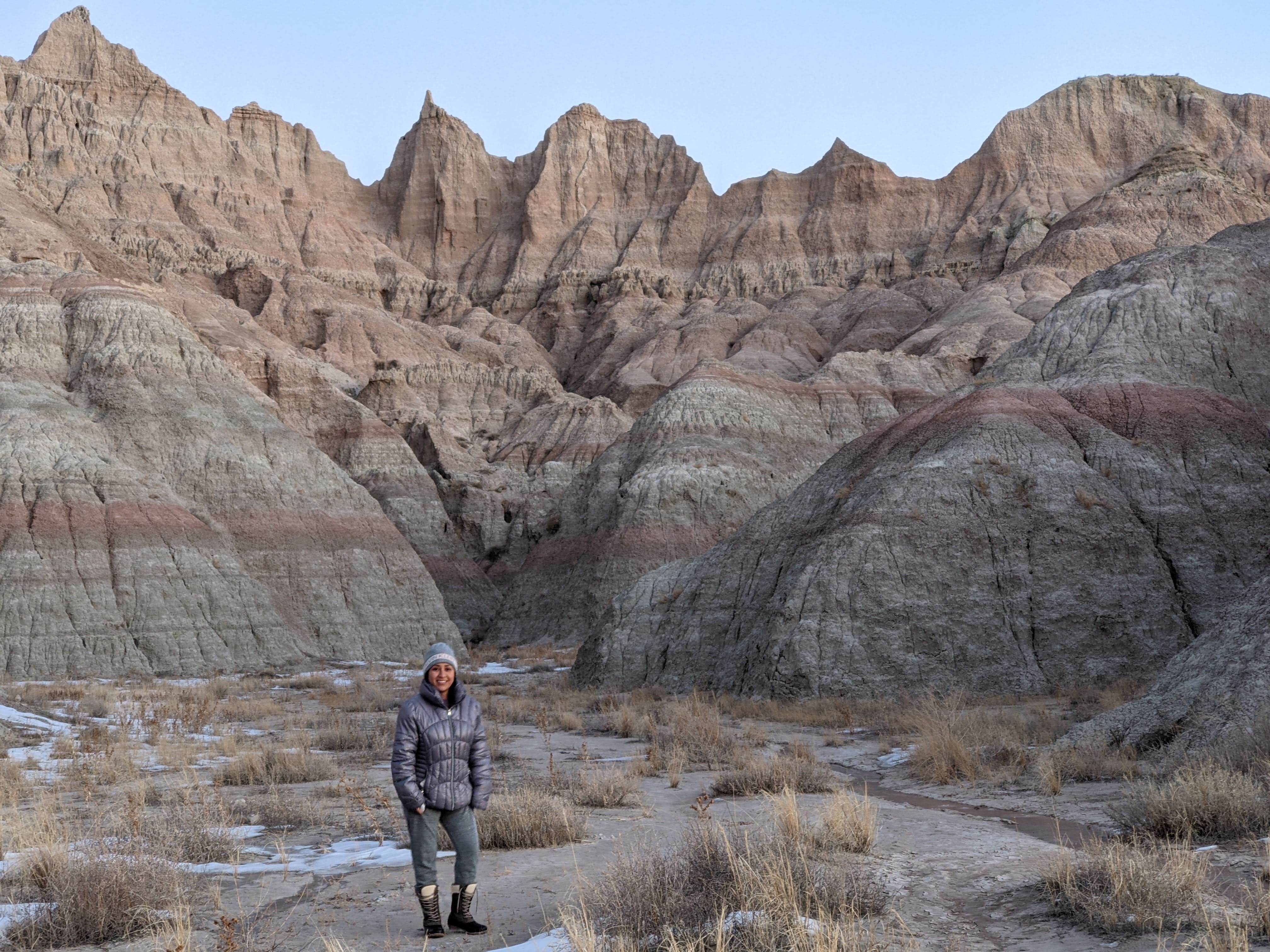 A person stands on a rugged trail surrounded by snow patches and dry grass in Badlands National Park. The landscape features dramatic eroded rock formations with layered colors and jagged peaks under a clear blue sky.