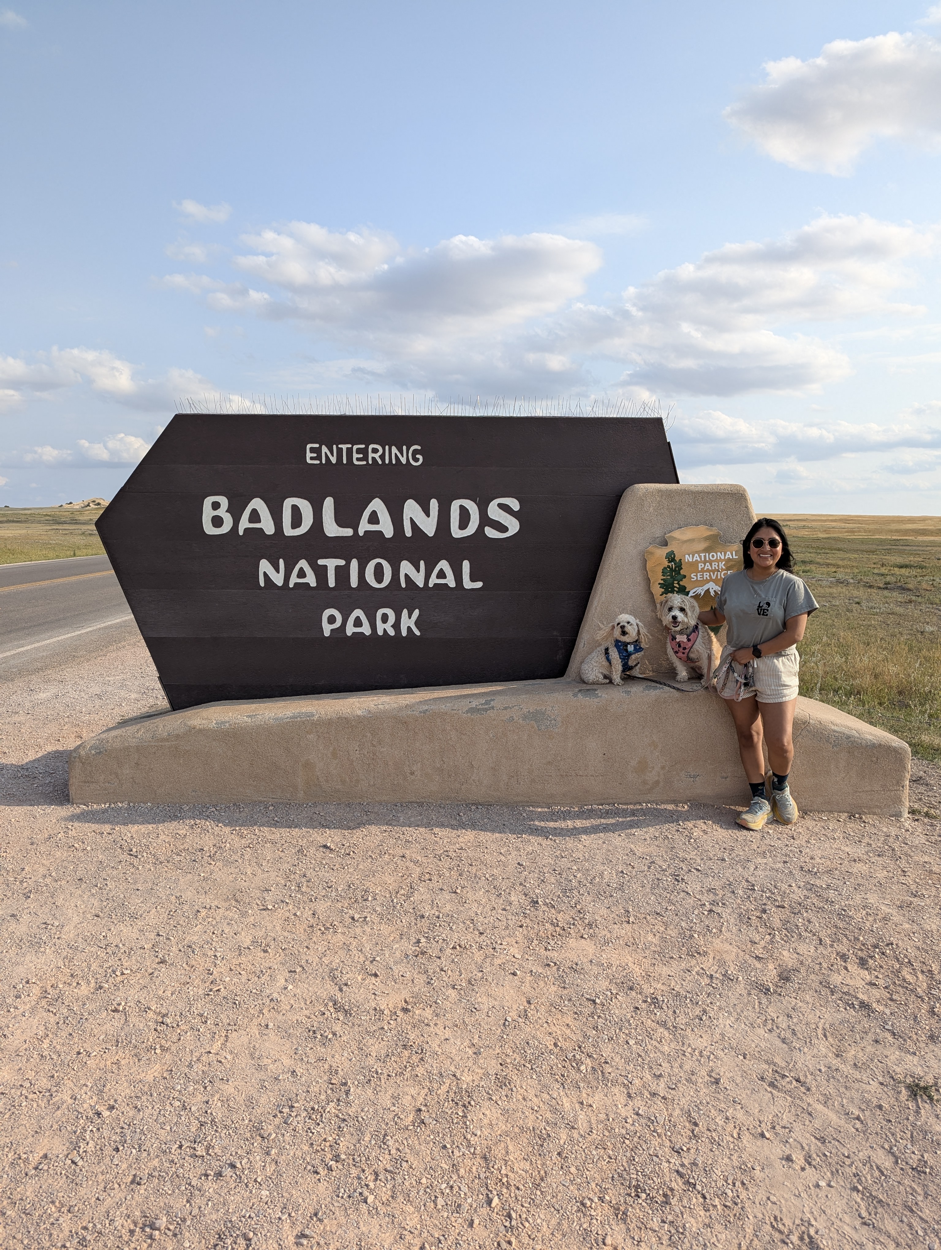 A woman wearing sunglasses and casual hiking clothes poses with two small dogs in front of the large entrance sign of Badlands National Park. The Brown sign features white lettering and the National Park Service emblem, set against an open prairie landscape under a bright blue sky