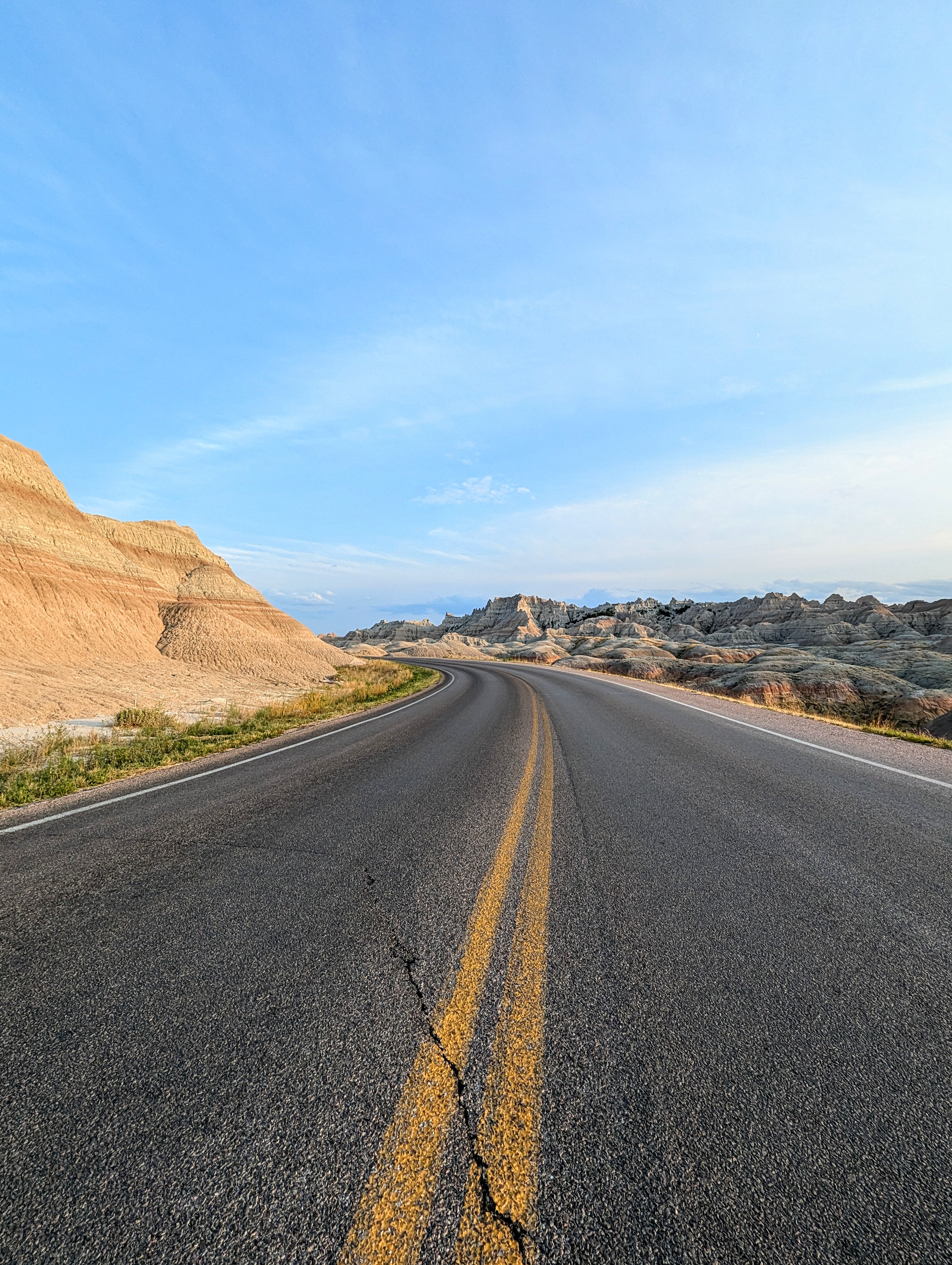 A winding asphalt road with double yellow lines curves through the rugged terrain of Badlands National Park, framed by sharply eroded rock formations and layered buttes. The early morning or late afternoon light casts soft shadows across the landscape under a wide, pale blue sky.