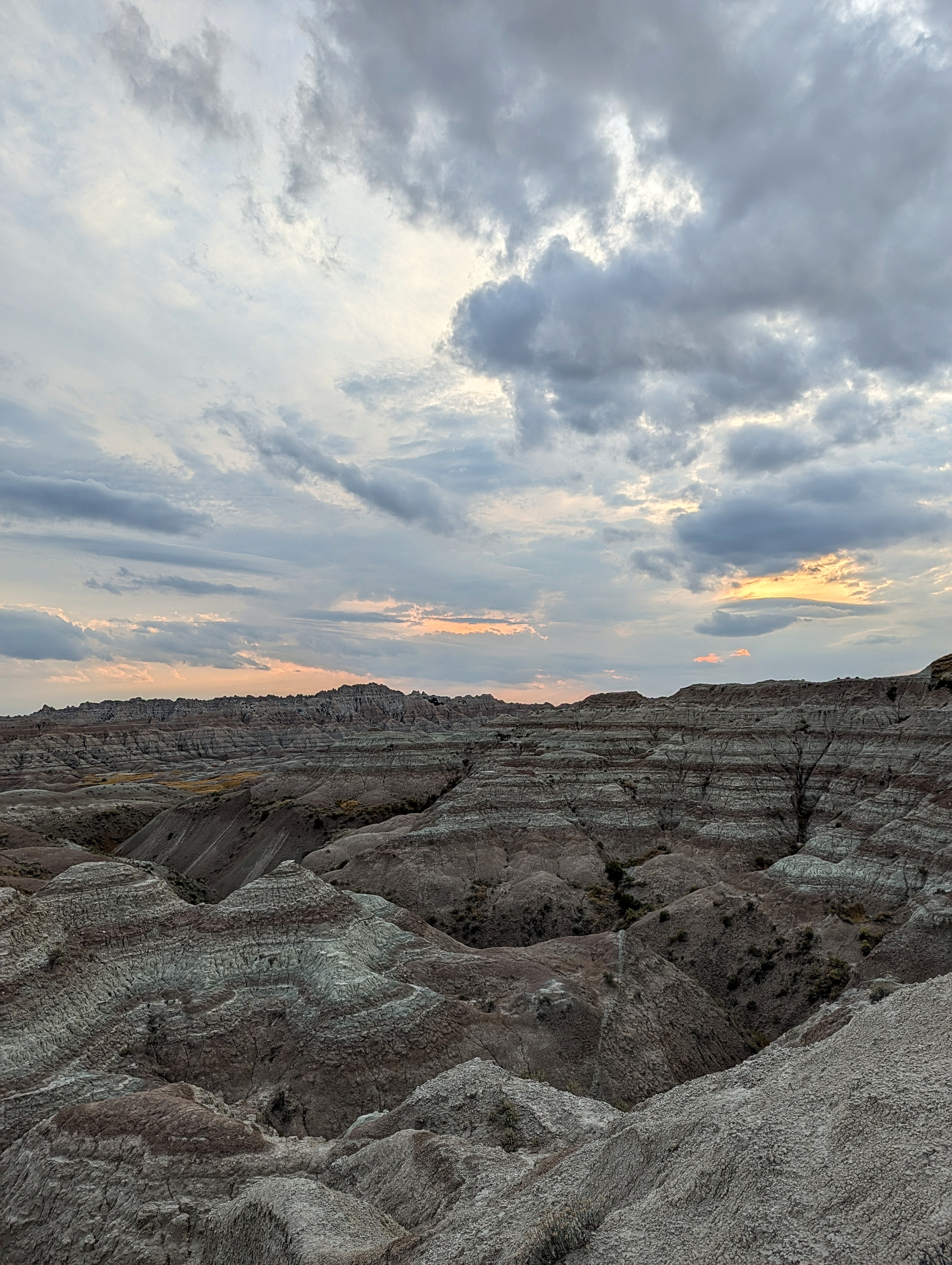 Dramatic layered rock formations stretch across the landscape in Badlands National Park under a moody, cloud-filled sky at sunset. Subtle hints of orange and pink peek through the clouds, casting a soft glow over the rugged terrain.