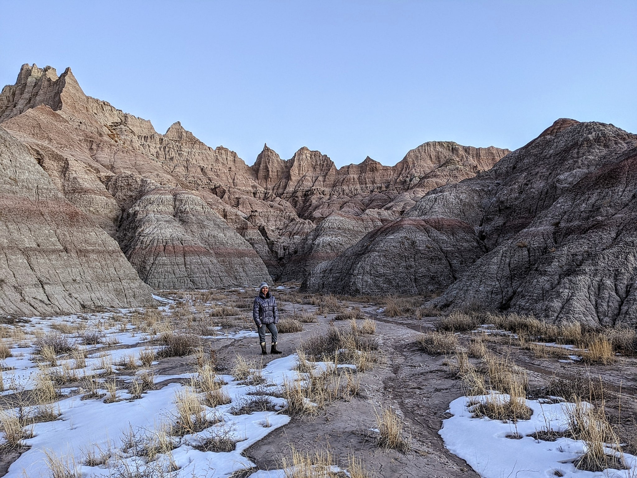 A person stands on a rugged trail surrounded by snow patches and dry grass in Badlands National Park. The landscape features dramatic eroded rock formations with layered colors and jagged peaks under a clear blue sky.