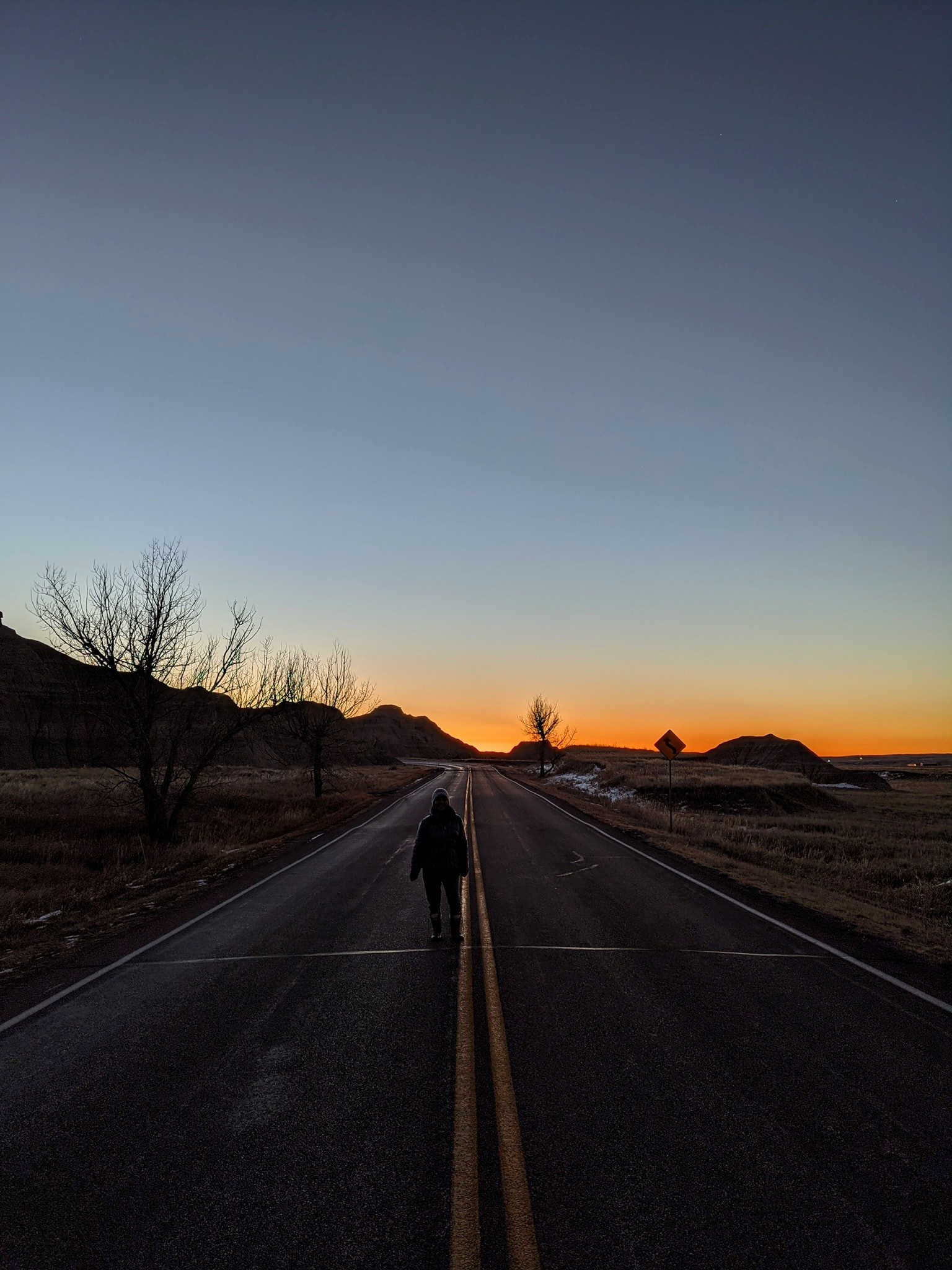 A person walks along a deserted road at sunrise in Badlands National Park. The sky transitions from a deep blue overhead to a warm orange glow near the horizon, silhouetting the surrounding hills and leafless trees. The peaceful, open landscape captures the quiet beauty of early morning in the park.